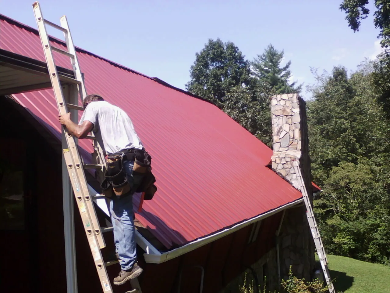 Roofer on a ladder beside a red metal roof and stone chimney — Soffit & Fascia Repair by Roofing Brothers in Trion